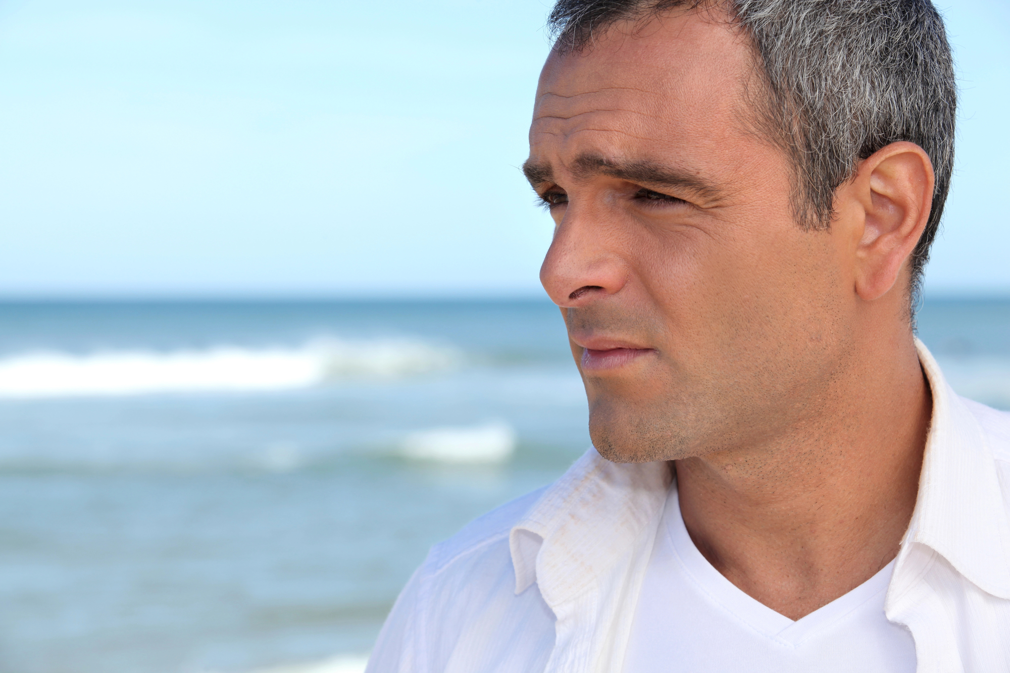 A man with short gray hair, wearing a white shirt over a white T-shirt, is standing on a beach. He is looking pensively into the distance with the sea and sky in the background.