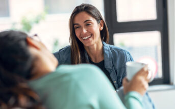 Two people sitting indoors, engaging in a relaxed conversation. One person with long brown hair in a denim shirt is smiling warmly at the other, who is holding a white mug. They are seated near a large window with blurred greenery outside.