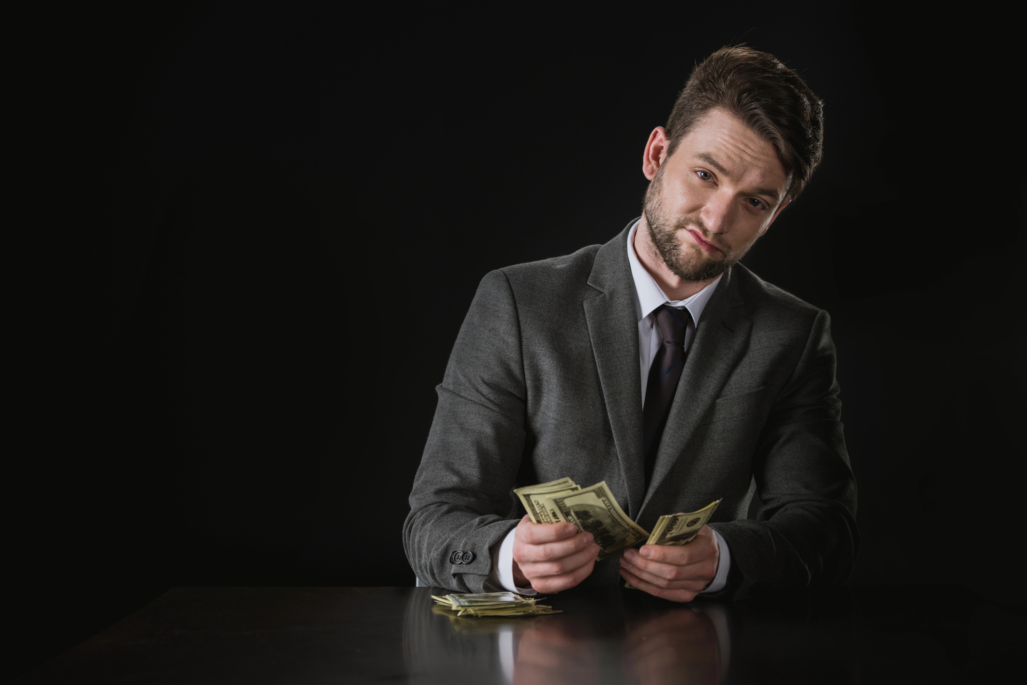 A man in a gray suit sits at a table in a dark room, counting a stack of US dollar bills. He has a serious expression and his head is tilted slightly. The table also has a small pile of more dollar bills. The background is black.