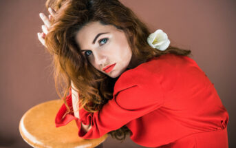 A woman with long red hair and a white flower tucked behind her ear is leaning on a wooden stool. She is wearing a red dress and gazing intently at the camera, her hands resting on top of the stool. The background is a warm, muted color.