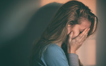 A woman with long hair, wearing a blue shirt, covers her face with her hands in an expression of distress. She appears to be emotional or upset, and the background is softly lit with a warm light.