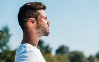 A man with short, dark hair and a beard stands outdoors in a profile view, facing left. He is wearing a white shirt and has a serene expression on his face. The background features blurred green trees and a clear blue sky.