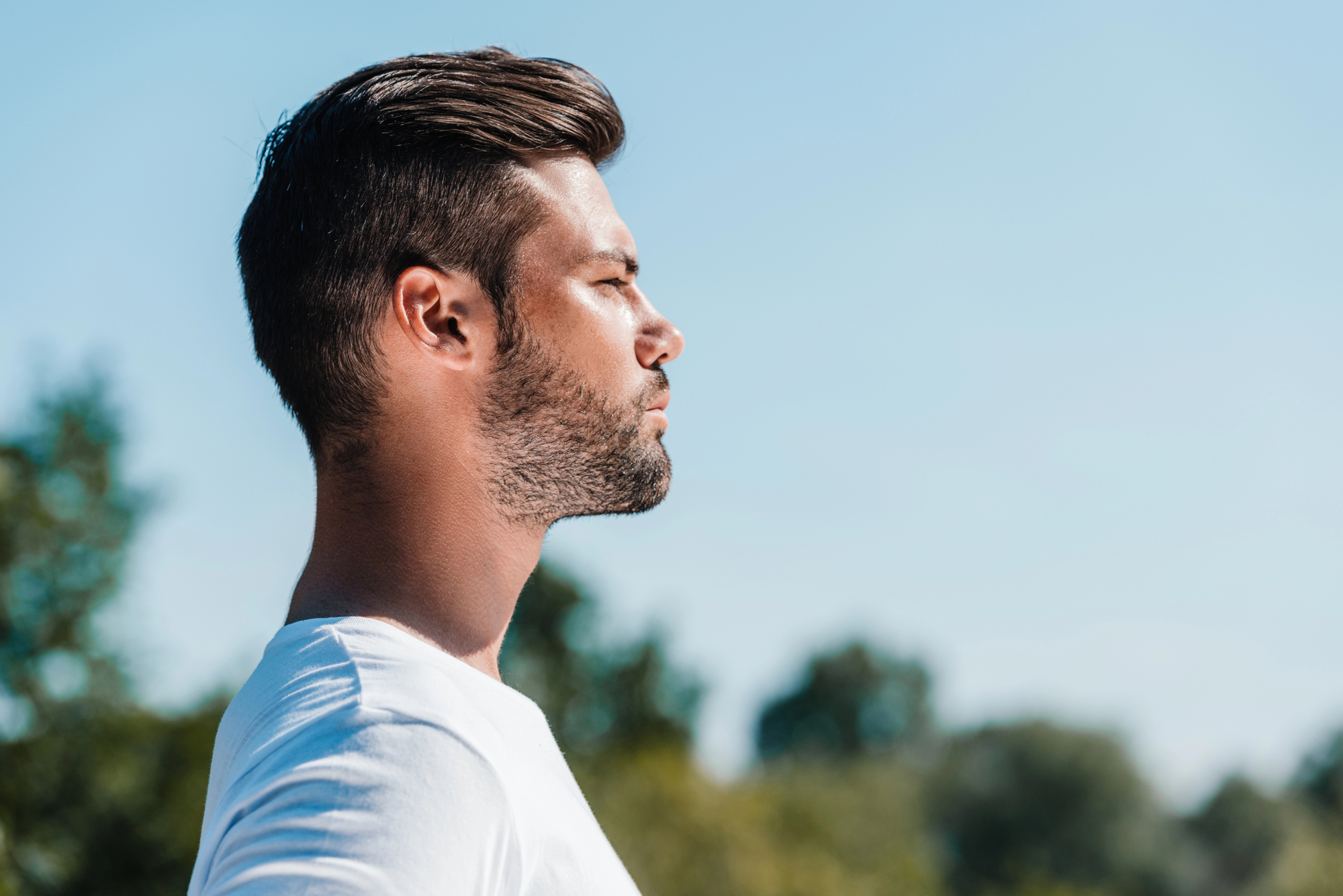 A man with short, dark hair and a beard stands outdoors in a profile view, facing left. He is wearing a white shirt and has a serene expression on his face. The background features blurred green trees and a clear blue sky.