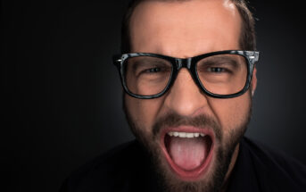 A close-up shot of a man with a beard and glasses, his mouth wide open as if shouting or expressing strong emotion. The background is dark, emphasizing his facial expression and the details of his face.