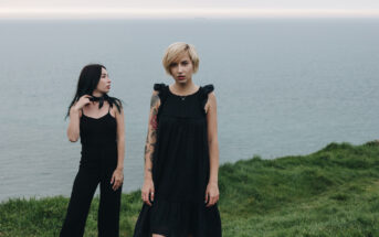 Two women stand on a grassy cliff overlooking a calm sea. The woman on the left, with long black hair, wears a black sleeveless top and pants, looking to her right. The woman on the right, with short blonde hair and a tattooed arm, wears a black dress, facing the camera.