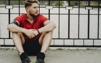 A man with short hair and a beard, wearing a red shirt and black shorts, sits on a paved ground with his knees up, leaning against a black metal fence with a body of water visible behind it. He has a fitness tracker on his wrist and an armband on his left arm.