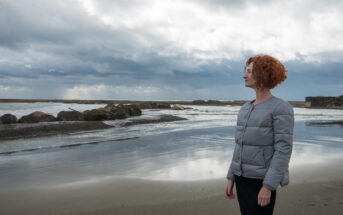 A person with curly red hair, wearing a gray jacket, stands on a sandy beach facing the ocean. The sky is cloudy and the sea appears calm with gentle waves. Rocks are visible in the water, and the overall scene has a serene atmosphere.