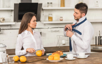 A woman is slicing oranges at a kitchen counter while looking at a man who is adjusting his tie. The counter is filled with breakfast items, including coffee cups, croissants, and oranges. The kitchen is modern with white shelves and a brick wall.