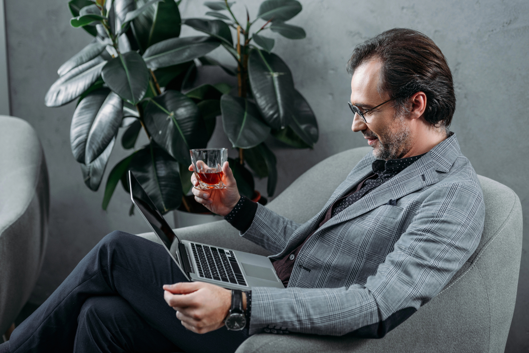 A man with glasses, wearing a gray checkered blazer and black pants, sits comfortably in a gray armchair. He smiles while looking at his laptop on his lap and holds a glass of a red beverage. A large green plant is in the background.