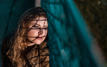 A woman with long, wavy hair is leaning against a teal wire fence. Shadows from the fence create lines across her face. She is looking to the side with a thoughtful expression. The background is blurred, drawing focus to her face and the fence.