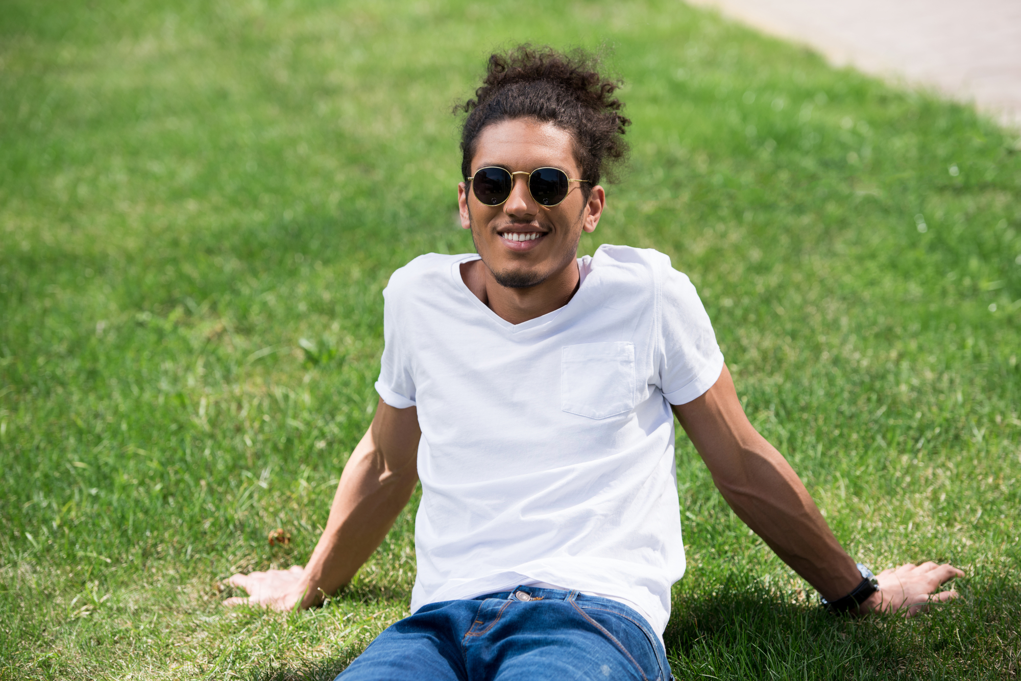 Young man with curly hair and sunglasses, wearing a white t-shirt and jeans, is sitting on the grass in a relaxed pose on a sunny day. He is smiling and leaning back on his hands with a carefree expression.