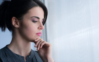 A woman with dark hair pulled back in a ponytail stands by a frosted window, looking out with a pensive expression. She rests her chin lightly on her hand, wearing a dark, long-sleeve top. The light from the window softly illuminates her face.