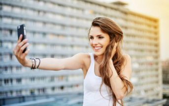 A young woman with long hair in a white tank top is smiling and taking a selfie with her smartphone. She is standing outside with a multi-story building in the background. The sunlight is softly illuminating the scene.