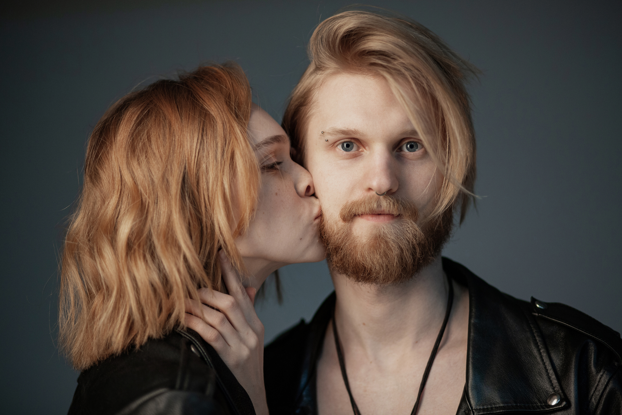 A person with short, wavy hair kisses the cheek of another person with a beard and longer hair. Both are wearing black leather jackets. The kissed person looks directly at the camera with a neutral expression, while the kisser's eyes are closed in the act.