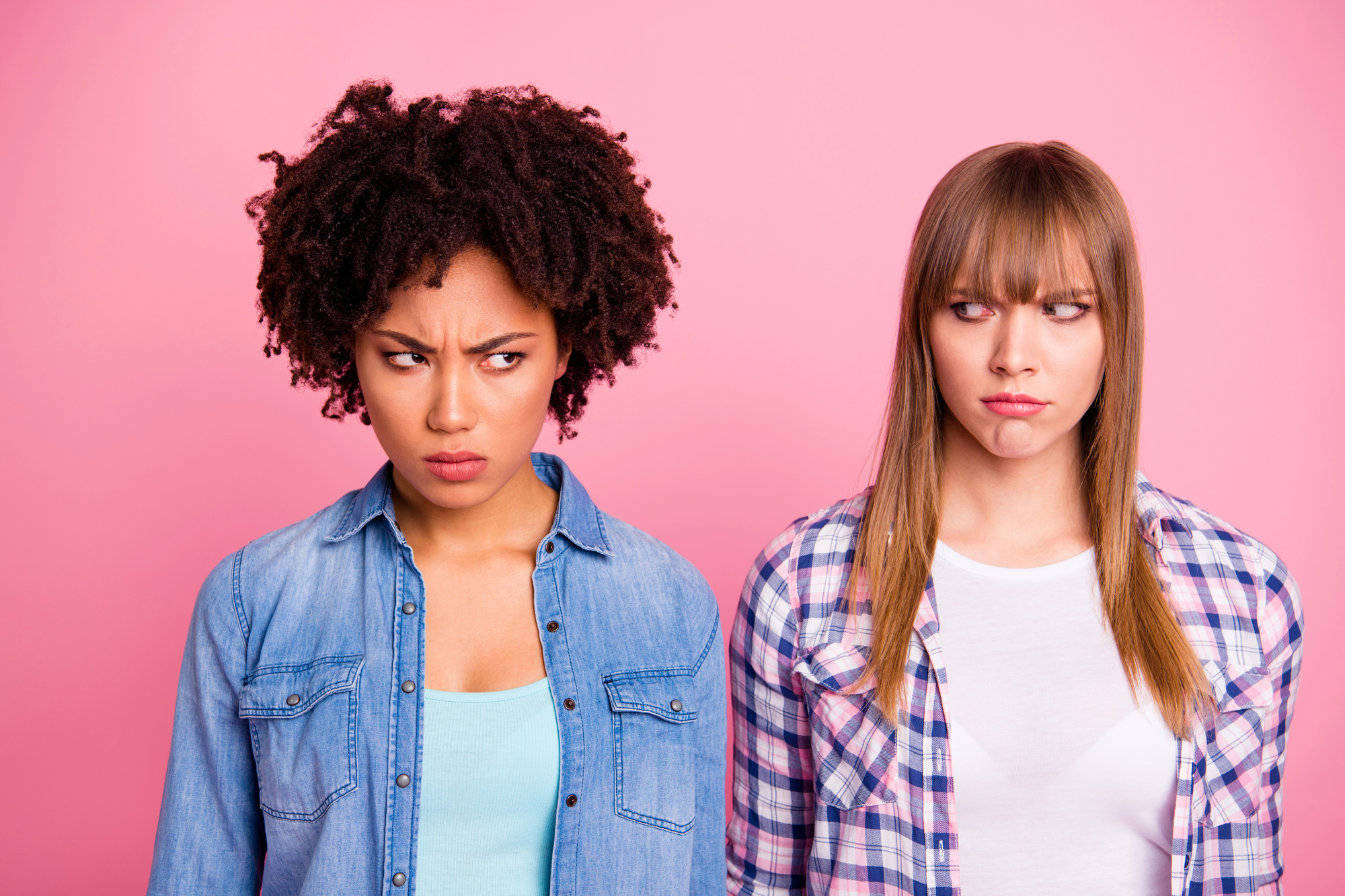 Two women stand against a pink background, looking at each other with suspicious expressions. The woman on the left has curly hair and wears a denim shirt over a light blue top, while the woman on the right has straight hair and wears a checkered shirt over a white top.