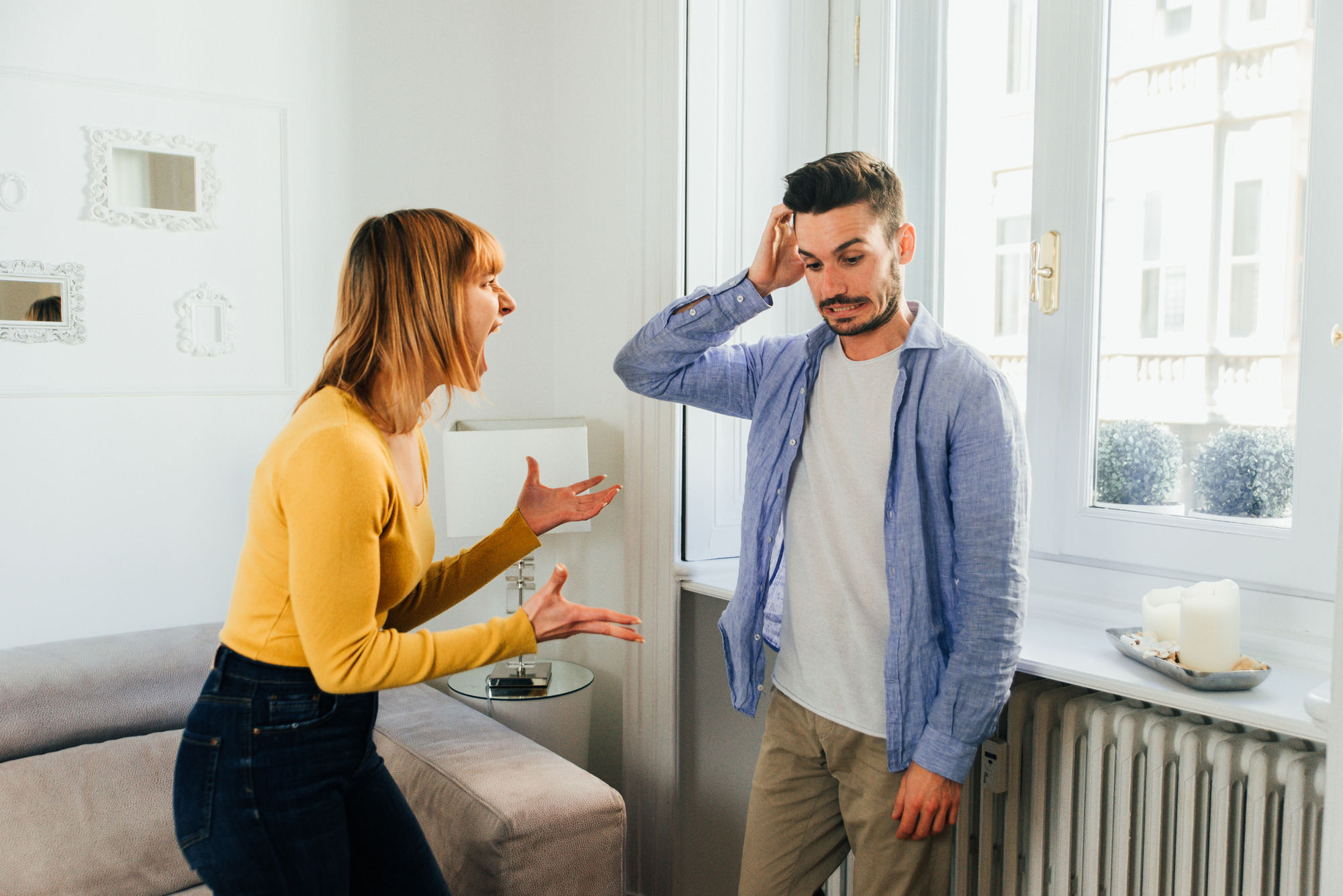 A woman with shoulder-length hair in a yellow sweater is yelling at a man with short hair in a blue shirt, who appears frustrated and is holding his hand to his head. They are standing in a bright room with white walls and large windows.