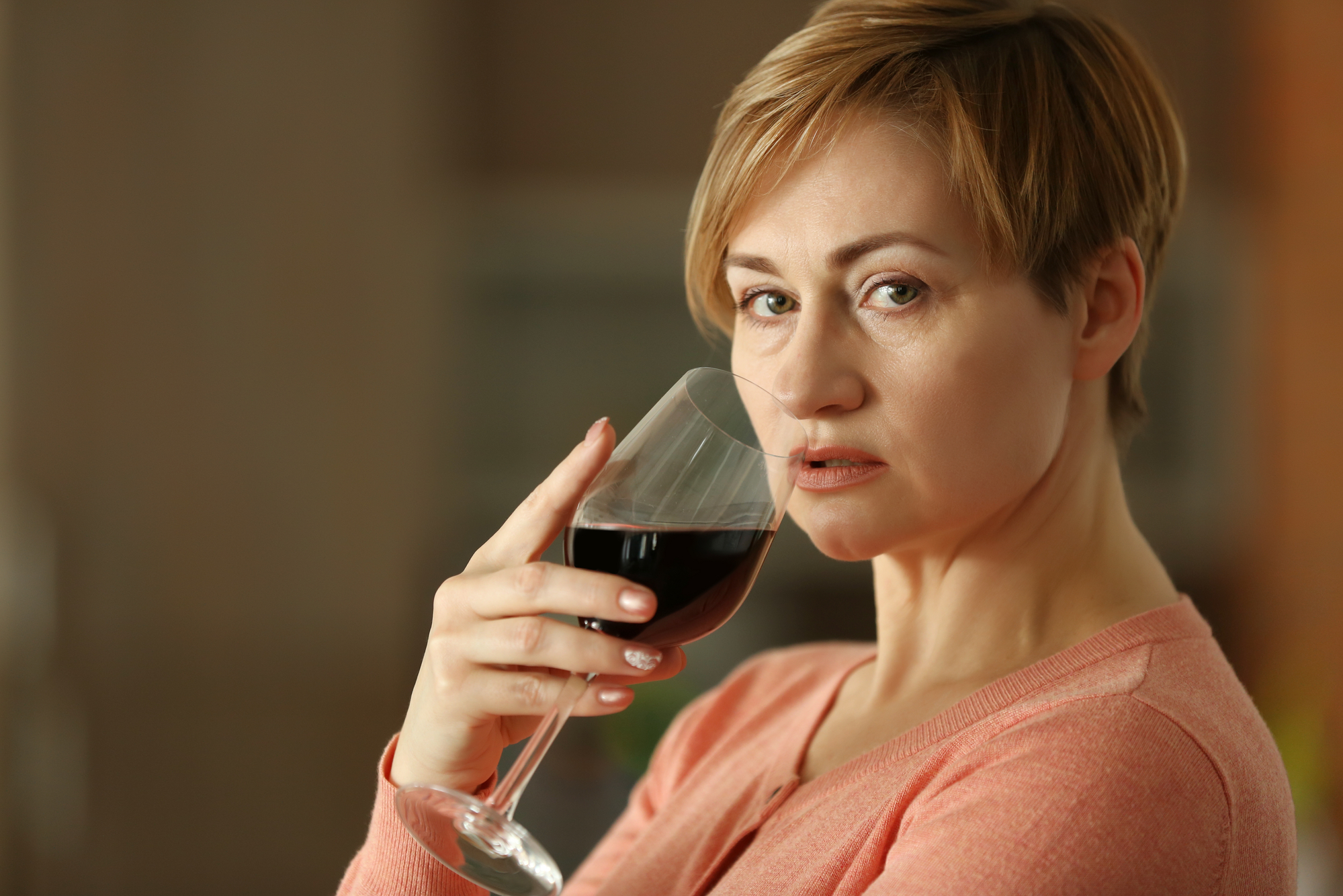 A person with short, light brown hair holds a glass of red wine close to their face, gazing directly at the camera. They are wearing a peach-colored top and stand against a blurred background. The expression is neutral, creating a contemplative mood.