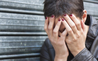 A person with short hair and red-painted nails rests their face in their hands, appearing distressed. They are wearing a dark leather jacket and are positioned in front of a corrugated metal wall.