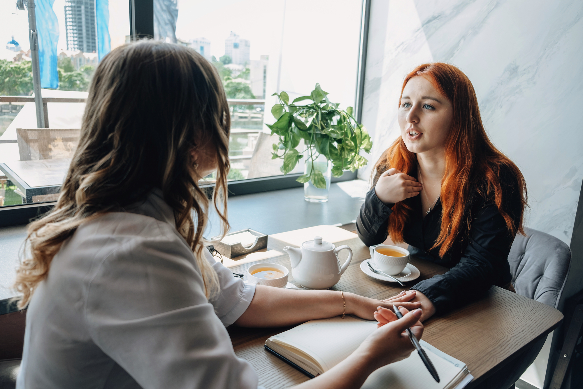 Two women sit across from each other at a table in a café, engaged in a conversation. One woman with red hair gestures while the other, writing in a notebook, listens. A teapot, cups of tea, and a plant are on the table, and a large window reveals an urban view outside.