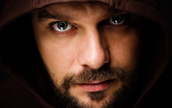 Close-up of a man with a brown hood covering his head, intensely gazing at the camera. He has a beard and mustache, with piercing eyes reflecting light. The backdrop is dark, highlighting his facial features.