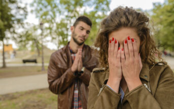 A woman with curly hair wearing a jacket covers her face with her hands while standing outdoors. Behind her, a man with short hair wearing a brown leather jacket and plaid shirt clasps his hands together as if pleading or apologizing.