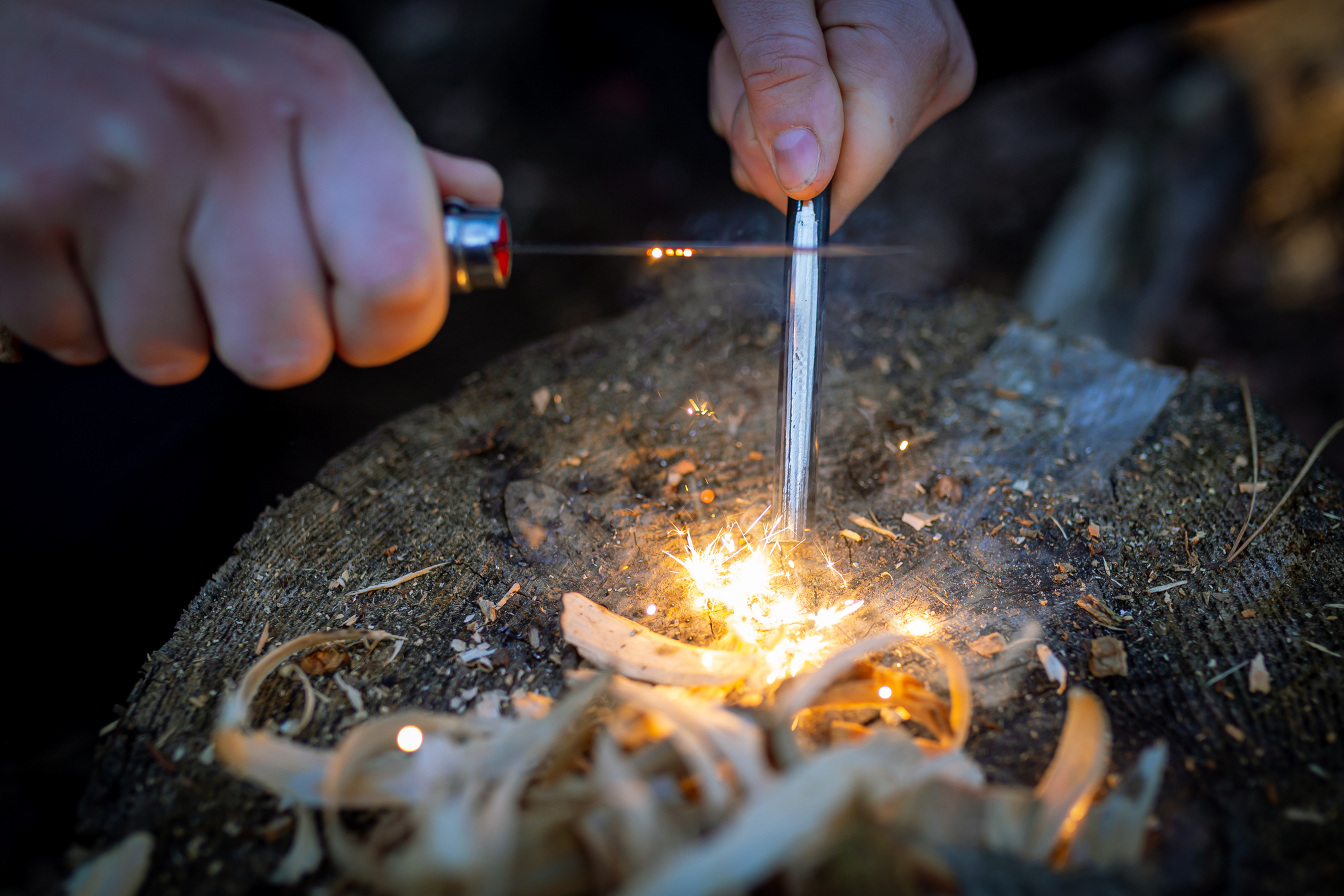 Close-up of hands using a ferrocerium rod and striker to create sparks and start a fire on a tree stump. Thin wood shavings and small pieces of kindling surround the spark.