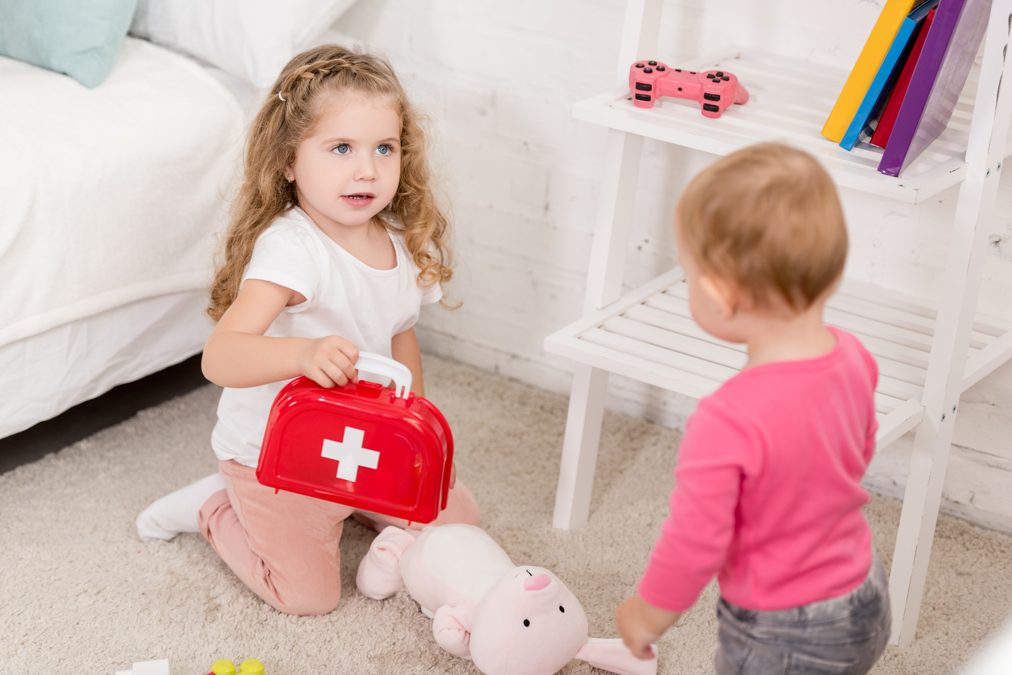 A young child with curly hair kneels on the carpet next to a plush bunny, holding a red toy medical kit. Another child, standing and facing away, watches. They are playing in a bright, tidy room with toys and books on a white shelf in the background.