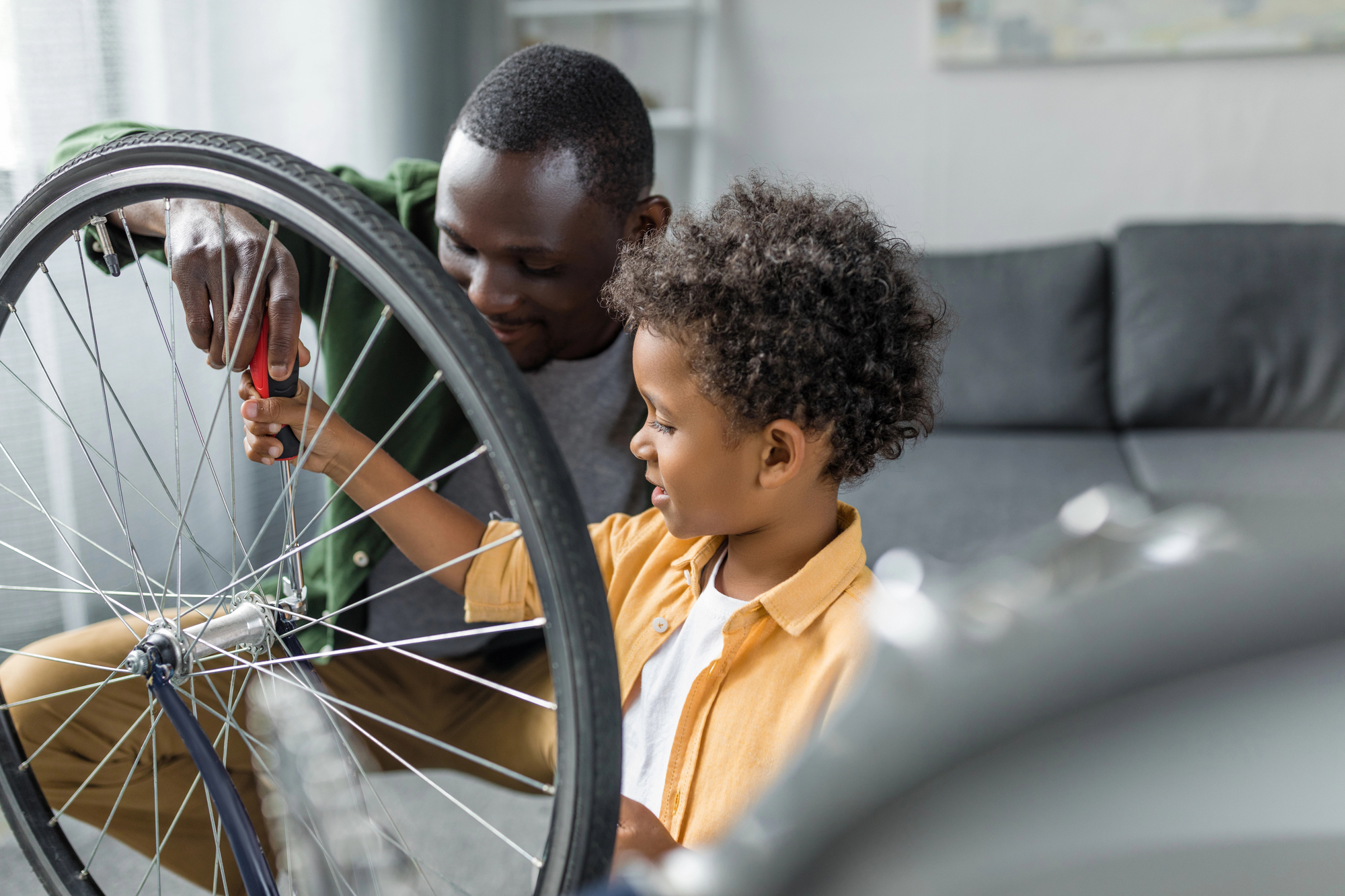 A man helps a young child repair a bicycle in a cozy living room. The man uses a screwdriver on the front wheel, while the child observes intently. The atmosphere is warm and collaborative, with a couch in the background.