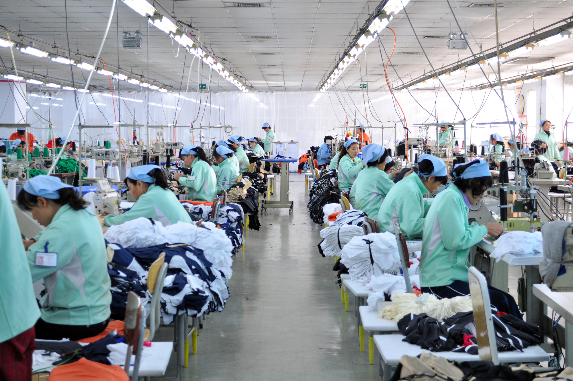 A large, brightly lit garment factory with rows of workers in green uniforms and blue caps, sitting at sewing machines and working on various clothing items. The factory has white walls, and tables are cluttered with fabrics and unfinished garments.