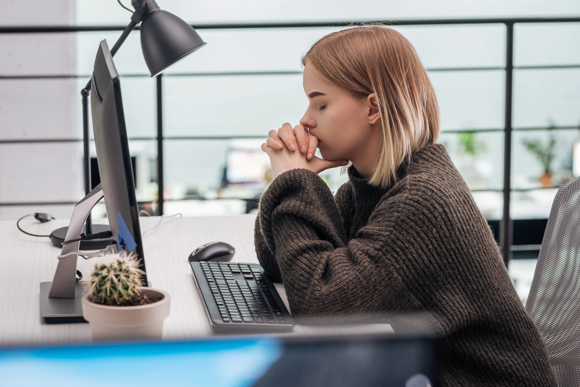 A person with shoulder-length blonde hair, wearing a brown sweater, sits at a desk in front of a computer. They are resting their head on their clasped hands, appearing deep in thought. There is a potted cactus and a desk lamp on the desk nearby.