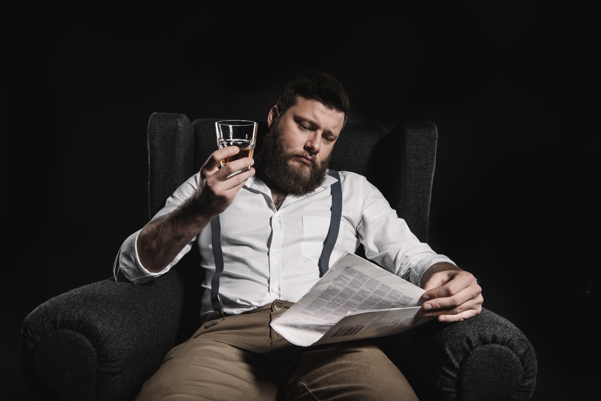 A bearded man with a serious expression sits in a dark gray armchair. He is wearing a white shirt with rolled-up sleeves and suspenders, holding a glass of whiskey in one hand and reading a newspaper with the other. The background is dark, highlighting him.