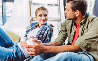 A young woman and man sit close to each other, each holding a white mug. The woman, dressed in a plaid shirt and jeans, smiles at the man, who wears a green shirt over a red T-shirt, both relaxed in a cozy indoor setting with light streaming in from a window.