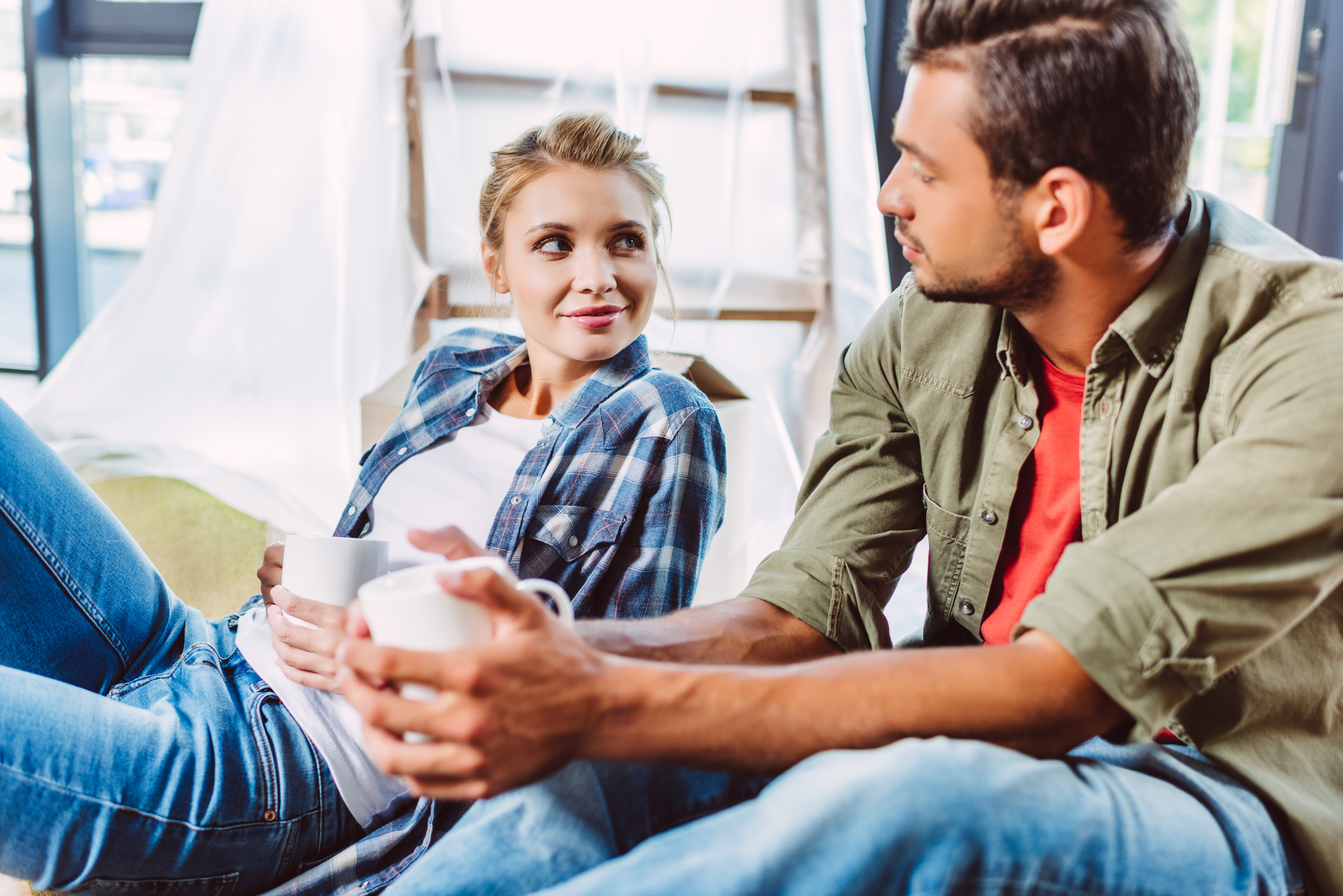 A young woman and man sit close to each other, each holding a white mug. The woman, dressed in a plaid shirt and jeans, smiles at the man, who wears a green shirt over a red T-shirt, both relaxed in a cozy indoor setting with light streaming in from a window.