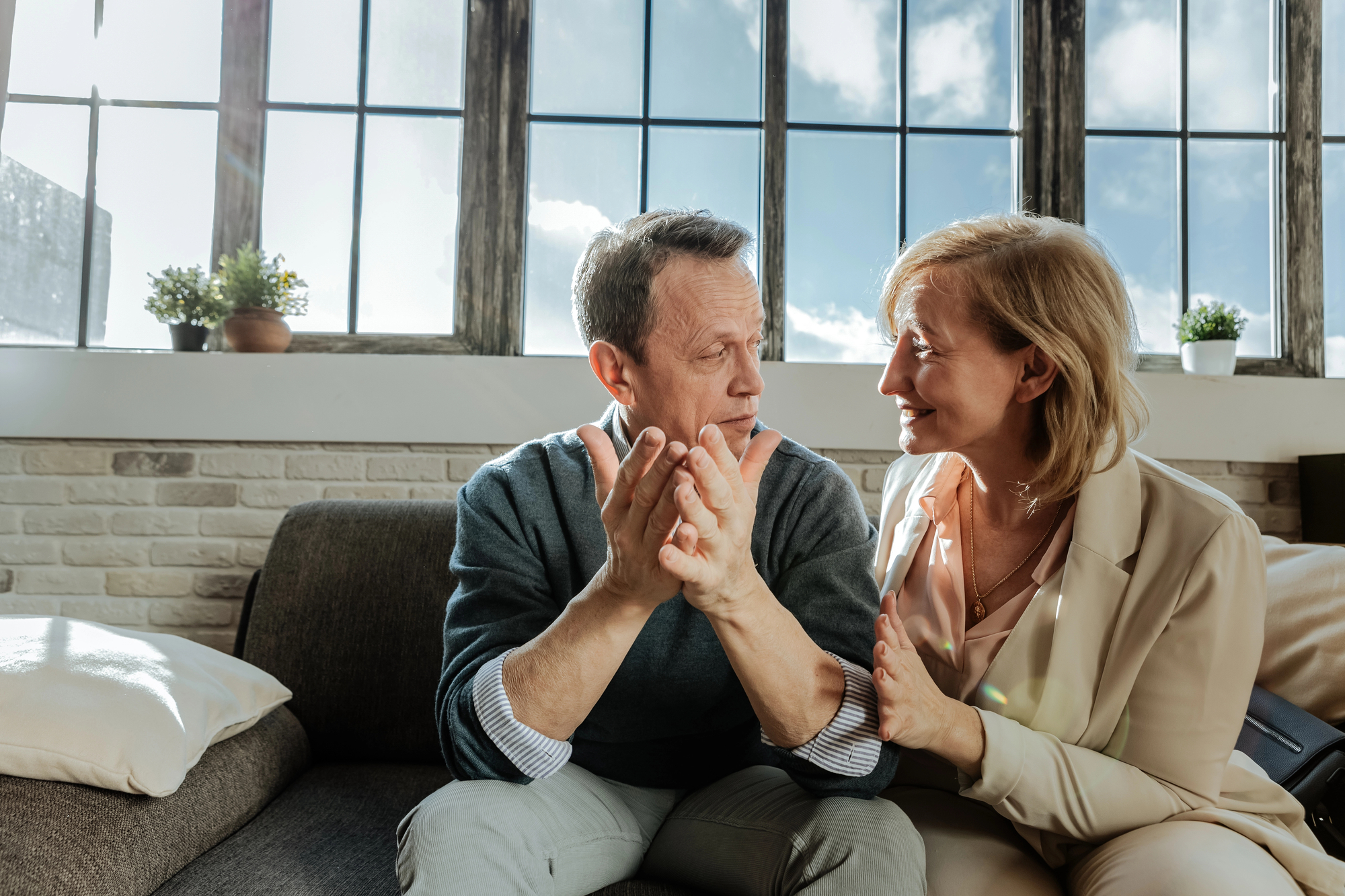 An older couple sits on a couch in a brightly lit room with large windows. They are holding hands and gazing at each other, smiling warmly. There are potted plants on the windowsill behind them.