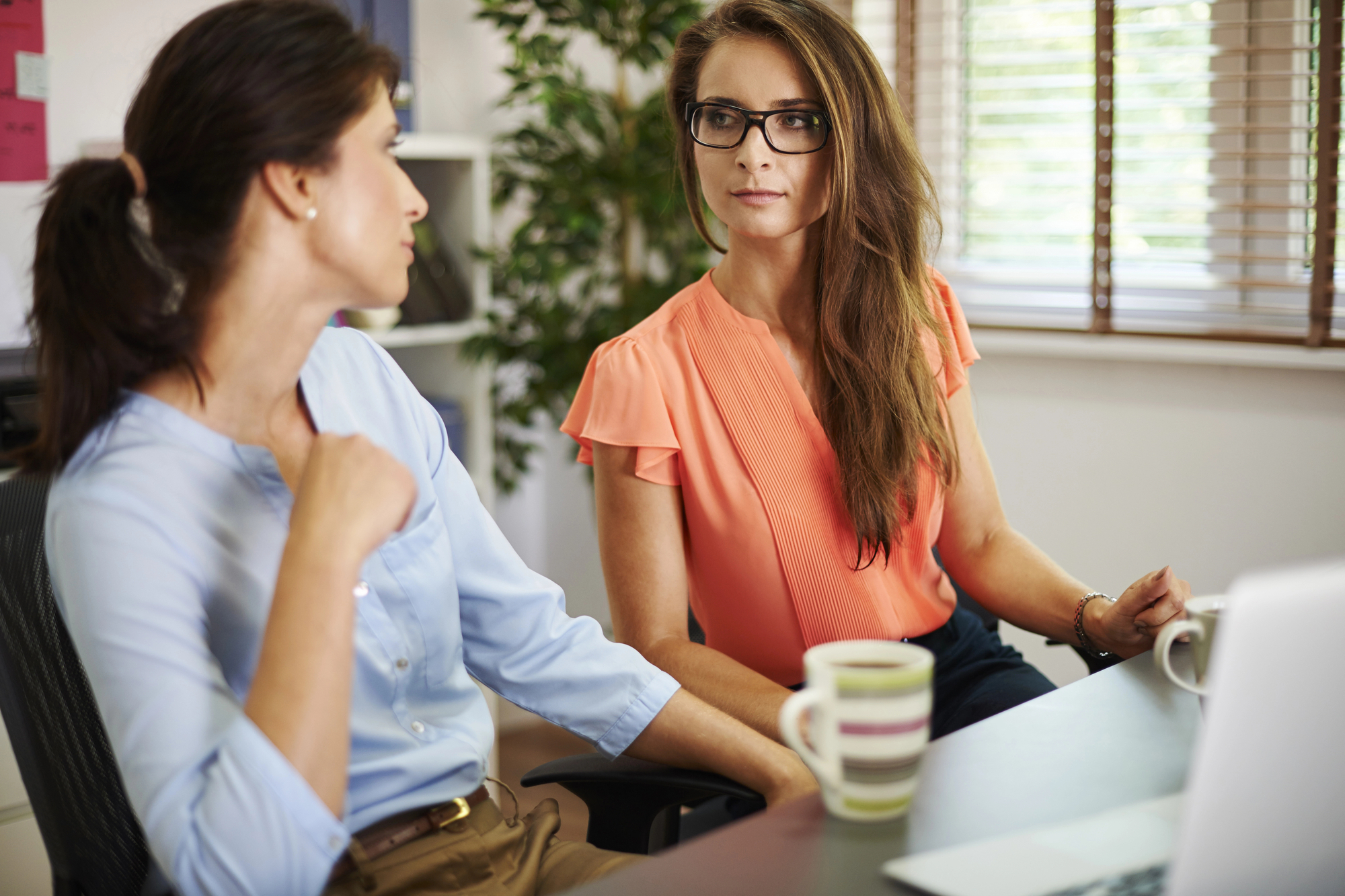 Two women are seated at a desk in an office, engaged in conversation. One, wearing a light blue shirt, is gesturing with her hand. The other, in an orange blouse and glasses, listens attentively. A laptop and two striped mugs are on the desk in front of them. Blinds cover the windows behind them.