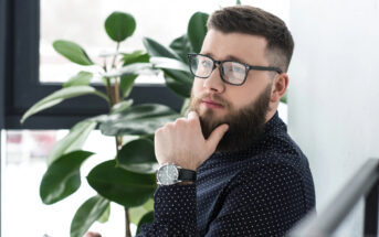 A bearded man with glasses, wearing a dark polka-dot shirt and a wristwatch, is sitting near a window with a potted plant behind him. He is looking thoughtfully into the distance, with one hand resting on his chin.