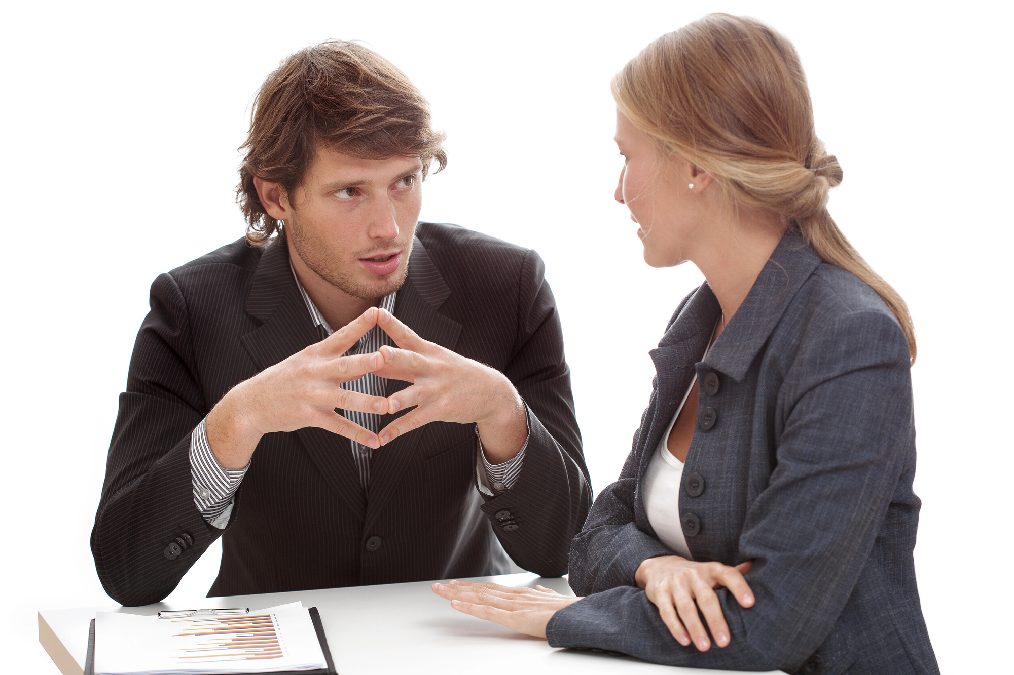 A man and a woman are sitting at a table engaged in a conversation. The man is gesturing with his hands, while the woman listens attentively. Both are dressed in business attire, and a document with charts is visible on the table in front of them.