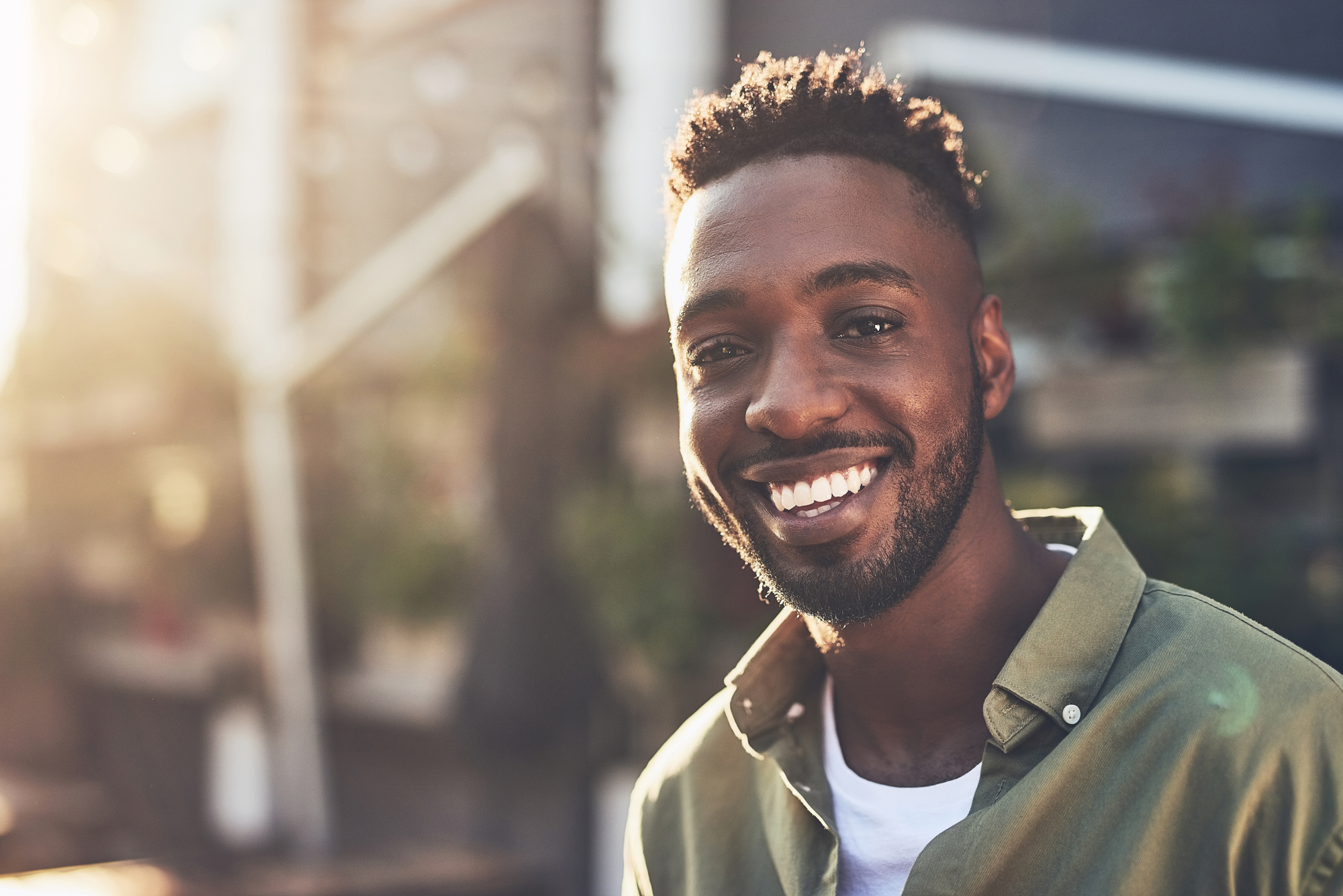 A man with a short beard and curly hair smiles warmly at the camera. He is wearing a green shirt over a white tee. The background is softly blurred, suggesting an outdoor setting with sunlight illuminating the scene.