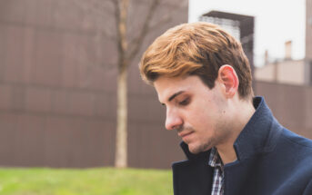 A young man with light brown hair and a short beard looks down with a thoughtful expression. He is wearing a navy blue coat and a checked shirt. The background features a blurred out-of-focus urban setting with a tree and a building.