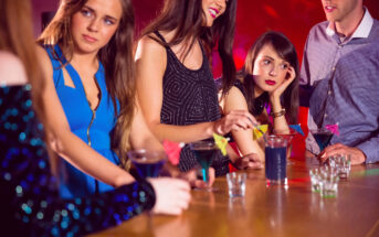 A group of young adults is gathered at a bar. Women in stylish dresses and a man in a button-up shirt are seen holding colorful cocktails with decorative paper umbrellas. One woman looks bored, resting her head on her hand, while others are engaged in conversation.