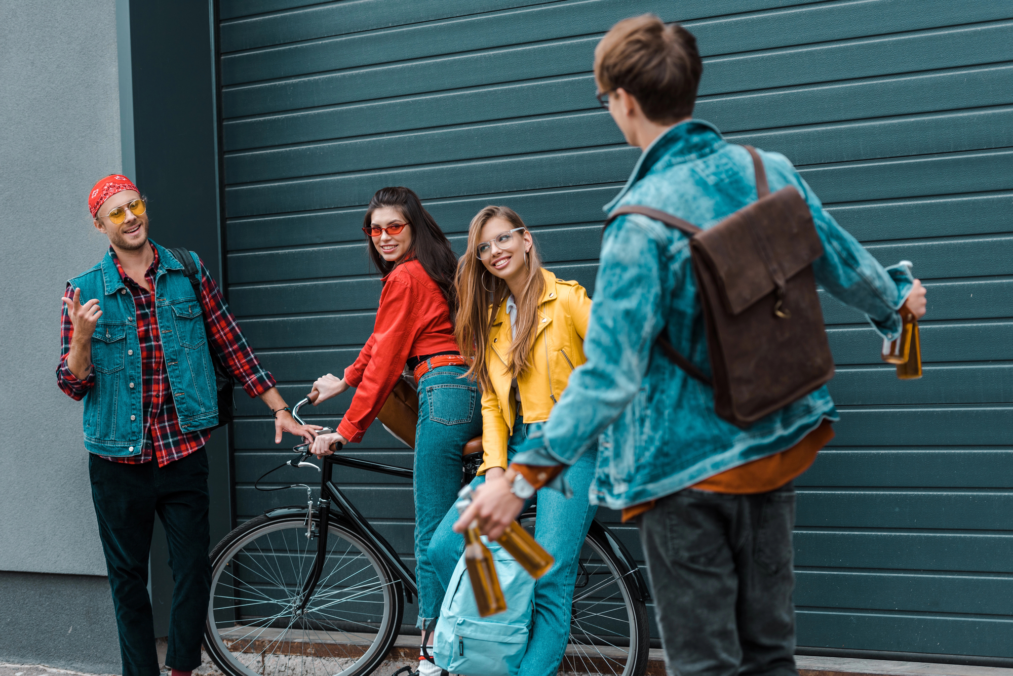 Four friends stand and chat near a closed garage door. Two women are leaning on a bicycle while smiling, and two men are holding beverage bottles, one of them with a backpack. They are dressed casually and appear to be enjoying a lighthearted conversation.