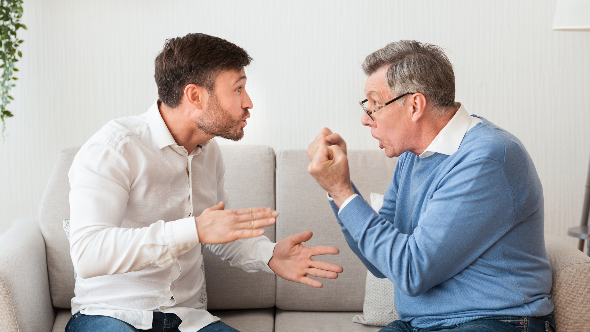 A younger man and an older man, both sitting on a light-colored sofa, are engaged in a heated discussion. The younger man, in a white shirt, gestures with his hands while the older man, in glasses and a blue sweater, responds animatedly with clenched fists.