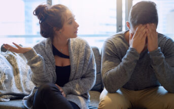 A woman gestures with frustration while sitting on a couch next to a man who is covering his face with his hands. The man appears to be distressed, and they are in a room with a large window in the background.