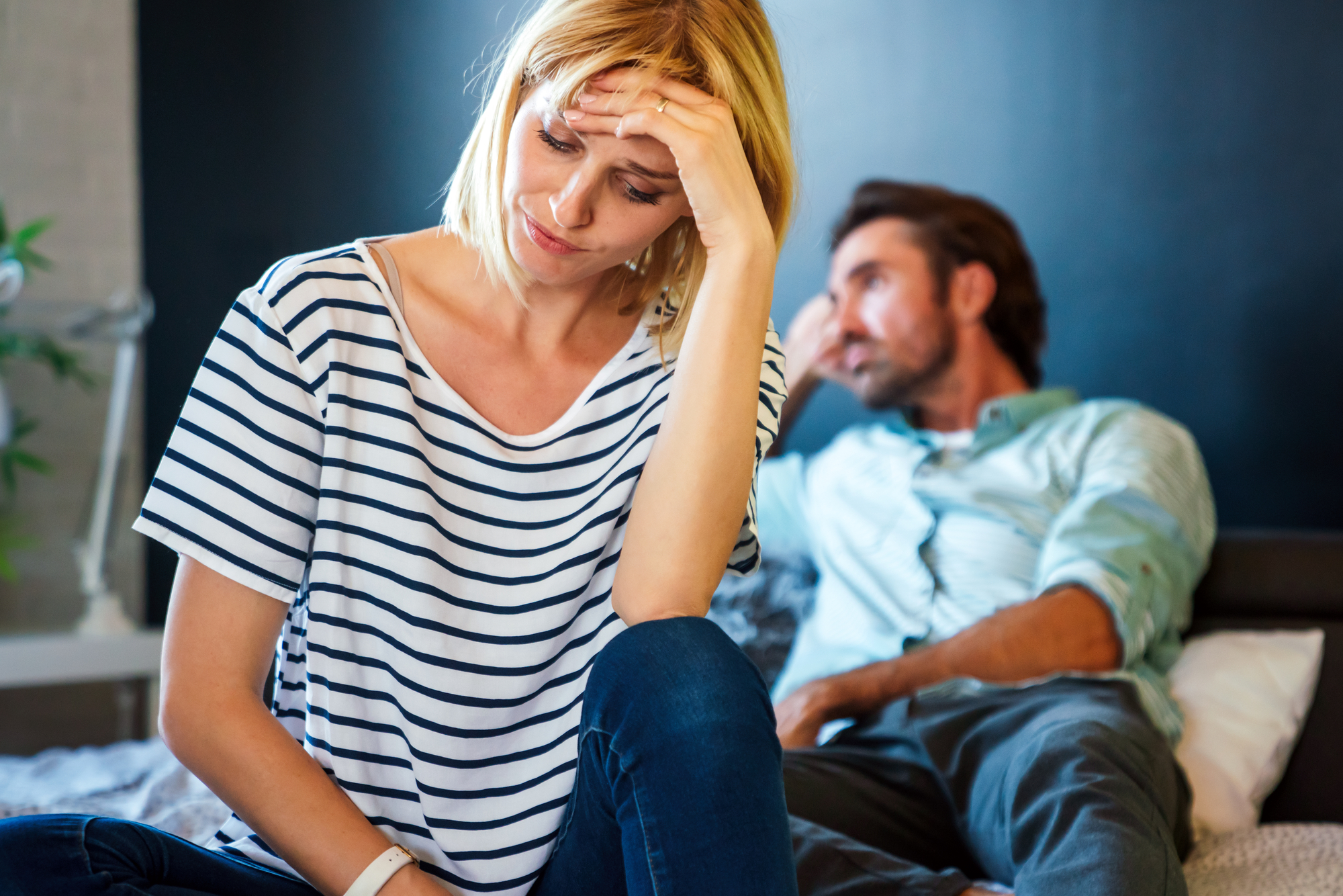 A woman with blonde hair wearing a striped shirt sits at the edge of a bed, looking distressed and holding her forehead. In the background, a man with dark hair leans back, propped up on the bed, appearing to be in a reflective or troubled state.