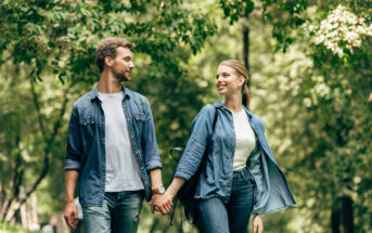 A couple holds hands while walking outdoors in a green, leafy park. Both are casually dressed in denim jackets and jeans. They are looking at each other and smiling, enjoying a sunny day together.