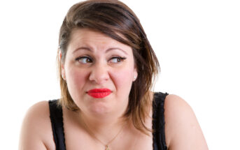 A woman with medium-length brown hair and red lipstick is making a disgusted or uncomfortable facial expression. She is wearing a black sleeveless top and a necklace with a small pendant. The background is plain white.