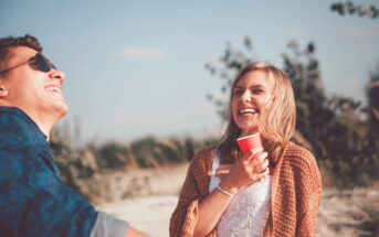 A man and woman enjoy a sunny day outdoors. The man is wearing sunglasses and smiling, while the woman, dressed in a light brown cardigan over a white top, holds a red cup and laughs. The background shows greenery and a clear blue sky.