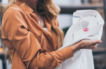 A woman holding a white shirt with a noticeable lipstick stain on the collar. She is wearing a brown shirt, and her long blonde hair is partially visible. The focus is on the lipstick stain as she examines the shirt.