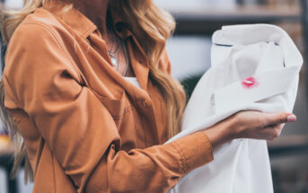 A woman holding a white shirt with a noticeable lipstick stain on the collar. She is wearing a brown shirt, and her long blonde hair is partially visible. The focus is on the lipstick stain as she examines the shirt.