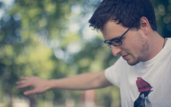 A young man with short dark hair and glasses stretches his arms out to the sides. He is wearing a white t-shirt with a graphic design. The background is blurred, featuring green foliage from trees. The image has a soft, serene ambiance.